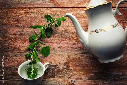 Tea time concept. White ceramic teapot, tea cup and fresh organic herbs, against wooden background.