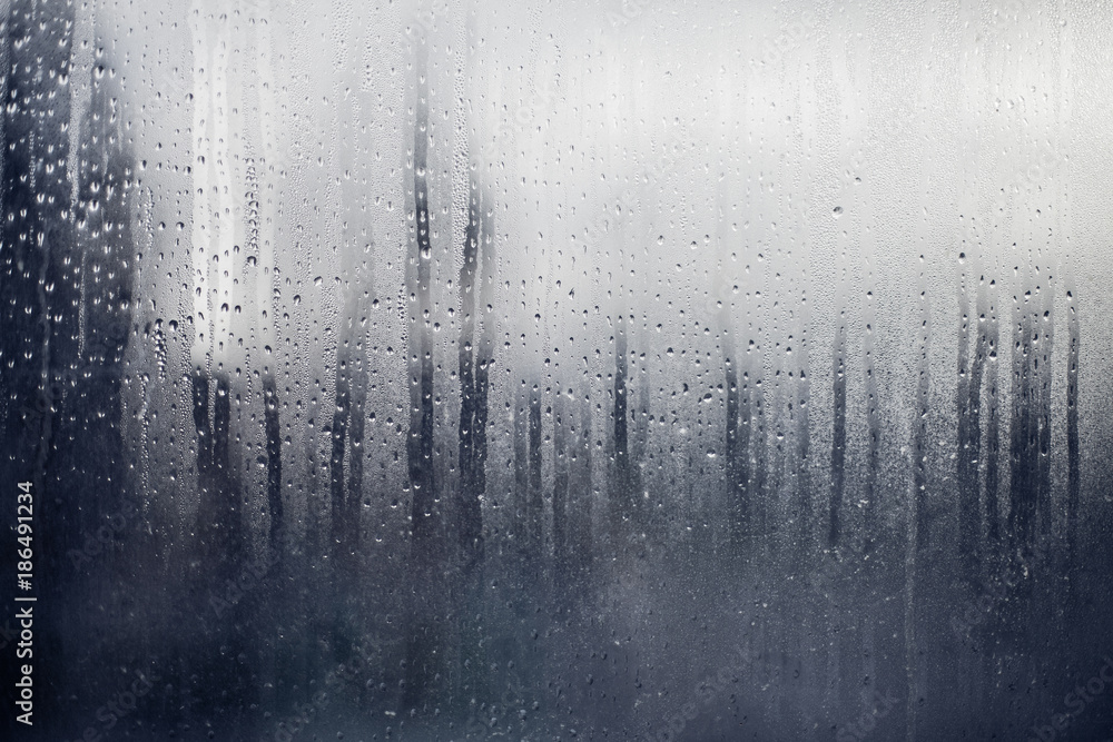 Water drops streaming on a window glass with grey city silhouette on background Water drops streaming on a window glass with grey city silhouette on background
