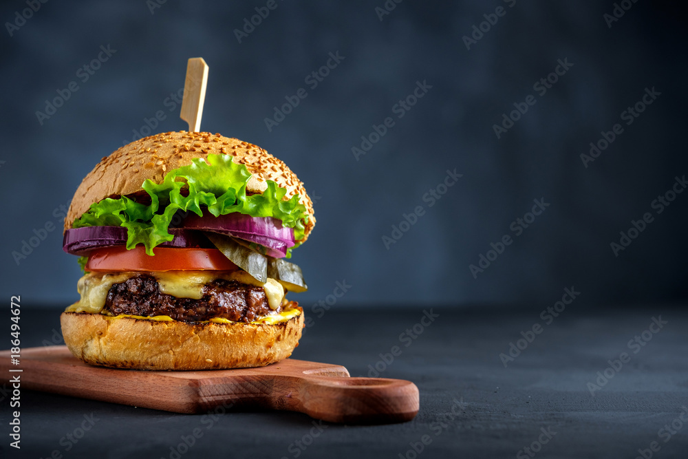 Tasty grilled beef burger with lettuce, cheese and onion served on cutting board on a black wooden table, with copyspace. - obrazy, fototapety, plakaty 