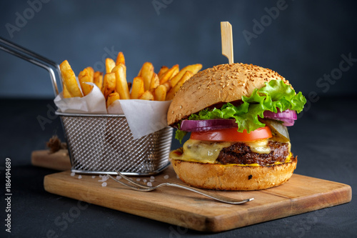 Tasty grilled beef burger with lettuce, cheese and onion served on cutting board with french fries on a black wooden table