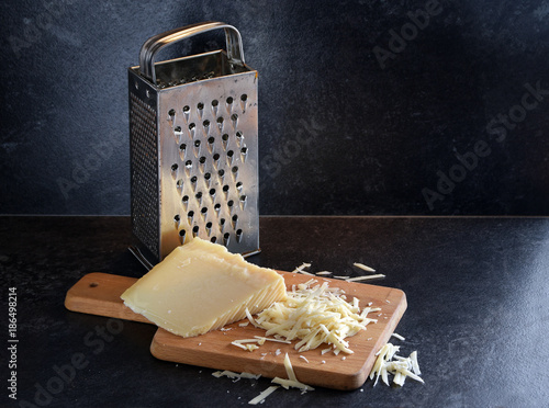 parmesan cheese on piece and grated and a used metal grater on a wooden cutting board against a dark slate background, copy space