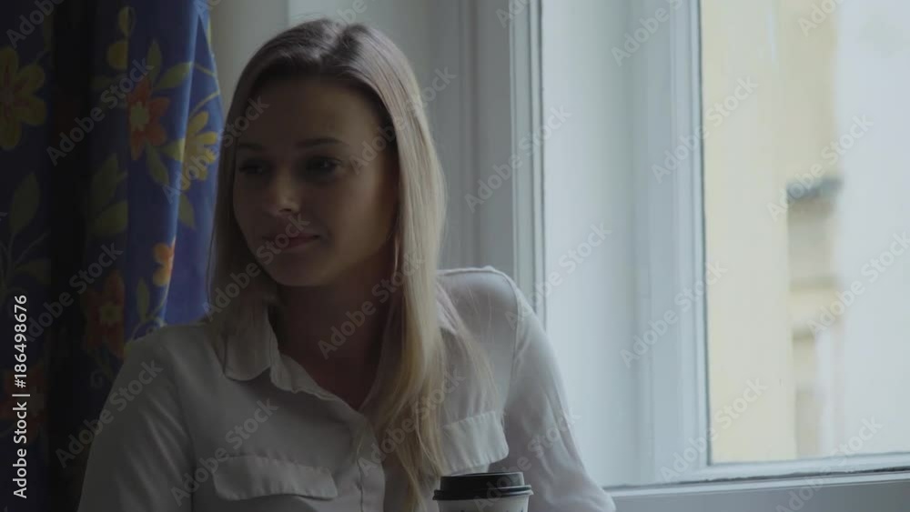 A young beautiful woman stands by a window, looks around the apartment and drinks coffee A young beautiful woman stands by a window, looks around the apartment and drinks coffee