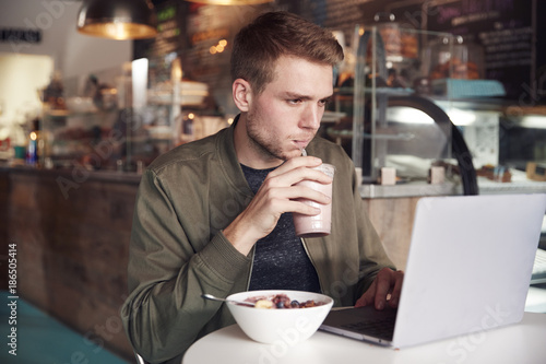 Young Man Using Laptop In Cafe Whilst Eating Breakfast