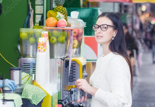 Photography Young attractive brunette girl in eyeglasses wearing white sweater buying fresh squeezed juice in street food booth
