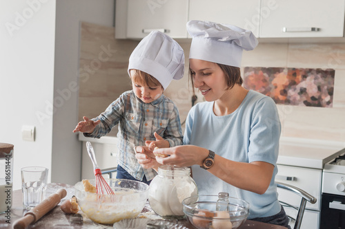 mother and adorable child in chef hats preparing dough at kitchen