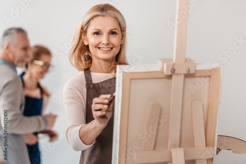Obraz na płótnie beautiful mature woman smiling at camera while painting on easel at art class