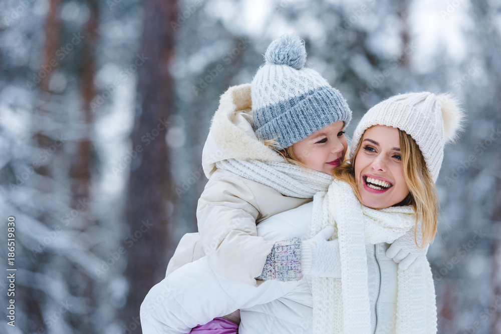 Fototapeta premium portrait of cheerful mother and little kid piggybacking together in winter park