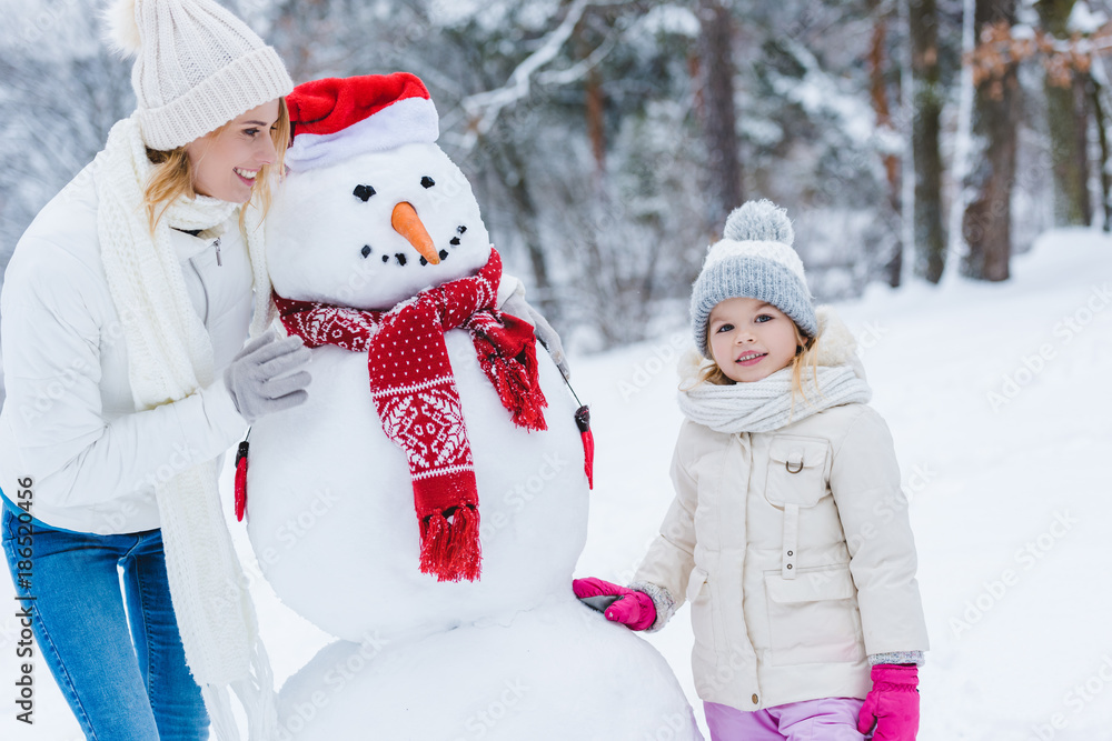 beautiful happy mother and daughter standing with snowman in winter park beautiful happy mother and daughter standing with snowman in winter park