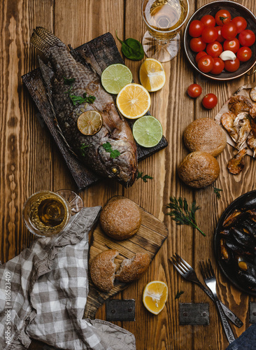 Top view of assorted seafood and baked fish with bread, tomatoes and white wine on wooden table