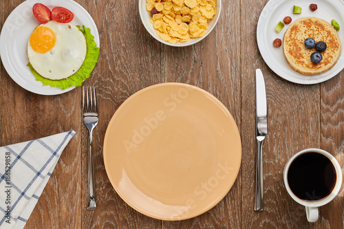top view of empty brown plate, pancake with blueberries and fried eggs with tomatoes and lettuce on a plates, cornflakes