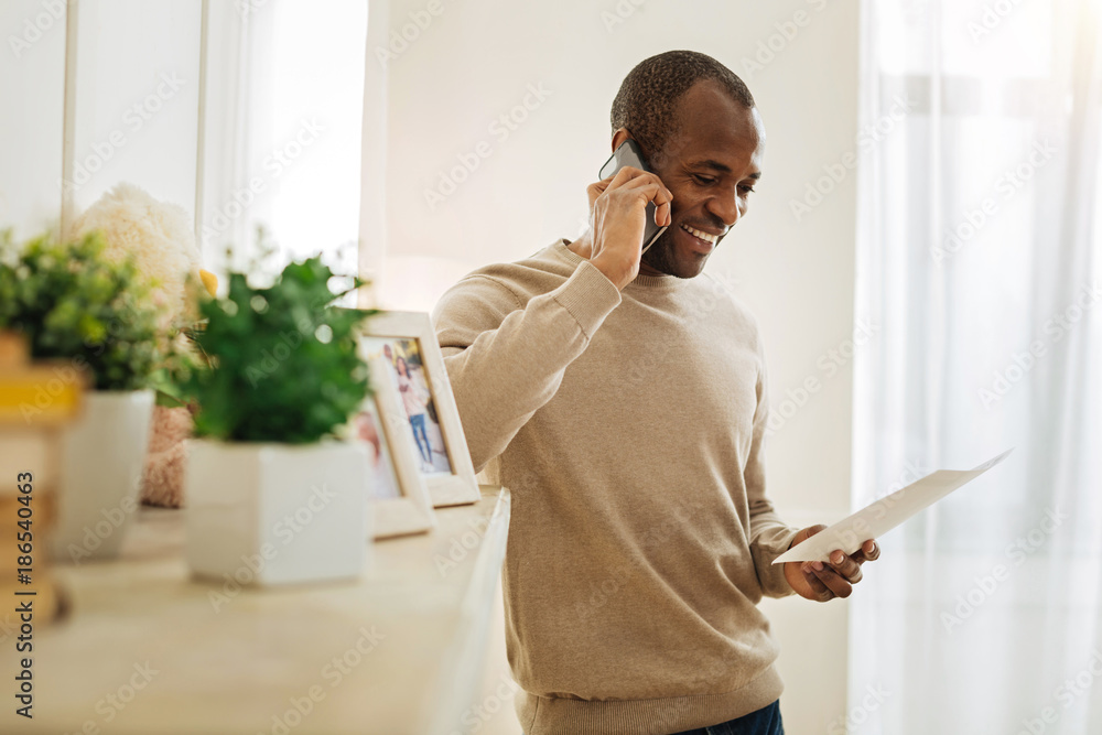 Pleasant talk. Vigorous bearded afro-american man laughing and talking on the phone while standing near the shelf with photos and flowers and looking at the sheet of paper Pleasant talk. Vigorous bearded afro-american man laughing and talking on the phone while standing near the shelf with photos and flowers and looking at the sheet of paper
