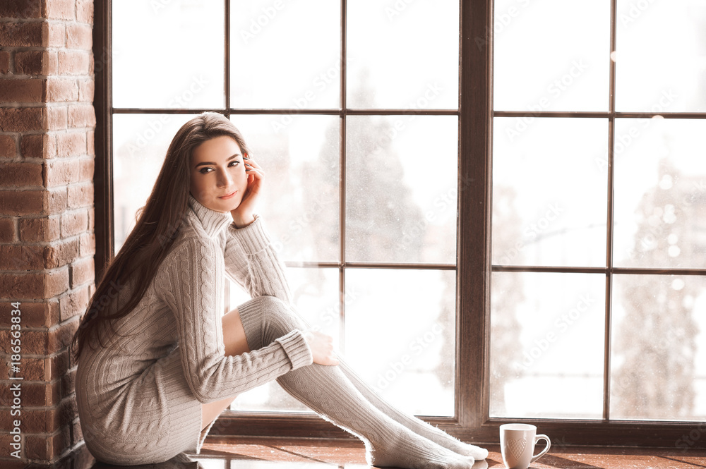 Beautiful smiling woman 24-26 year old sitting on windowsill wearing  knitted sweater and socks. Looking away. WInter season. 20s. Stock Photo