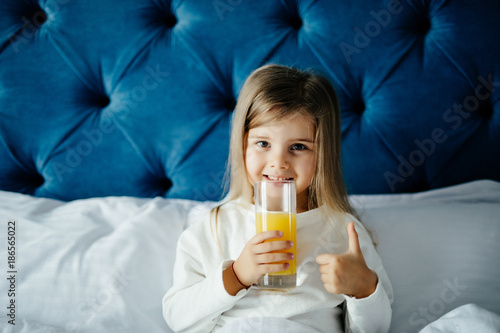 Beautiful girl girl holding glass of orange juice, showing thumb up gesture, looking at camera while sitting in bed