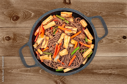 Delicious buckwheat noodles with tofu skin and vegetables in a cast iron wok on dark wooden background, top view