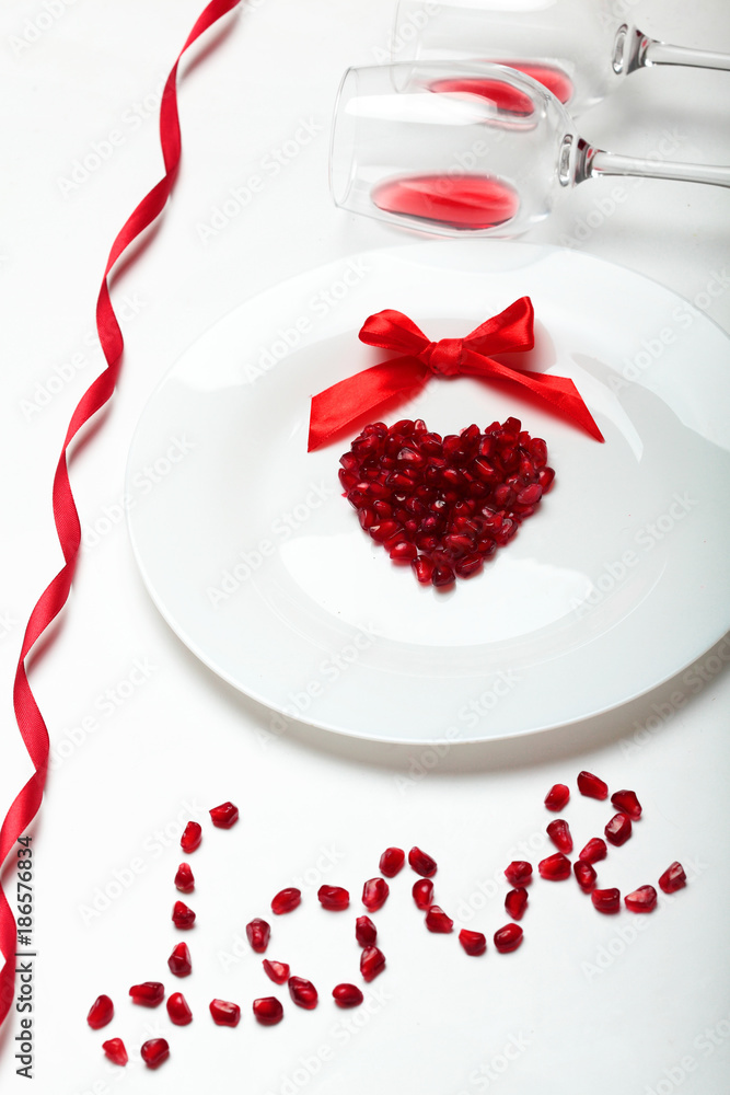 Pomegranate grains in a heart shape on white plate and red bow of ribbon two glass with wine. Word love of Pomegranate grains .On white background.Background for valentine's day.Minimalism.Closeup Pomegranate grains in a heart shape on white plate and red bow of ribbon two glass with wine. Word love of Pomegranate grains .On white background.Background for valentine's day.Minimalism.Closeup