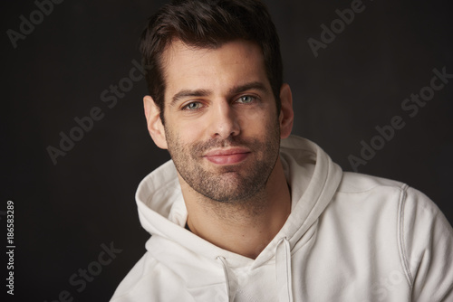 Studio shot of a handsome young man sitting at dark background while looking at camera and smiling.