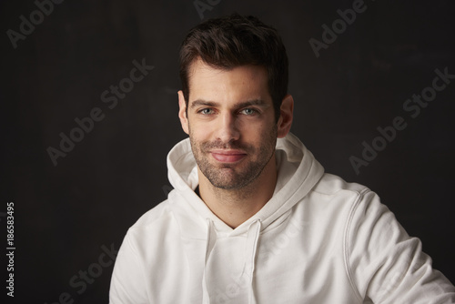 Studio shot of a handsome young man sitting at dark background while looking at camera and smiling.