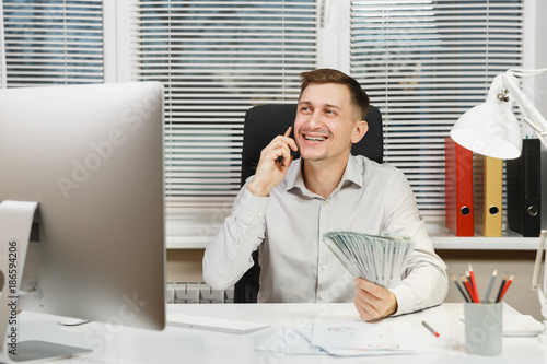 Handsome smiling business man in shirt sitting at the desk talking on mobile phone with lot of cash money, working at computer with modern monitor, lamp, documents in light office. Manager or worker. © ViDi Studio