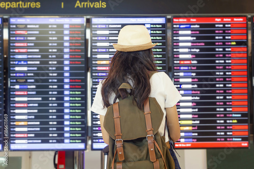 Young woman with backpack and carry on luggage in international airport, near flight information board.