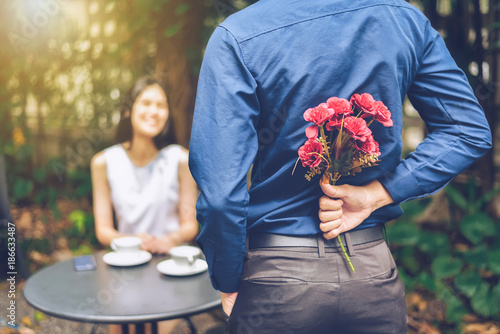The man is hiding red flowers behind him in order to surprise his girlfriend.
