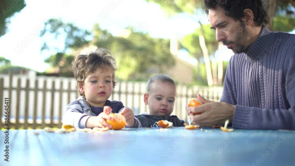 A father with two little sons peeling and eating tangerines in the garden A father with two little sons peeling and eating tangerines in the garden