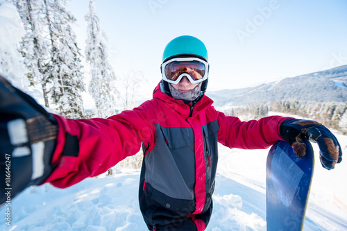 Shot of a man snowboarder taking a selfie, wearing helmet, skiing mask and colorful winter snowboard clothing, standing on top of the mountain at the ski resort