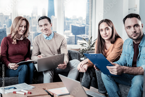Pleasant team. Friendly intelligent reliable workers sitting in the office on the sofa near the window looking straight and doing theirs tasks.