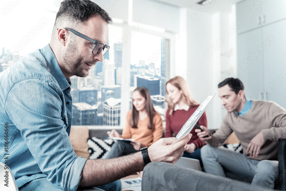 Obraz premium Interesting facts. Bespectacled thoughtful unshaken man sitting in the room on the chair holding the document and overlooking it.