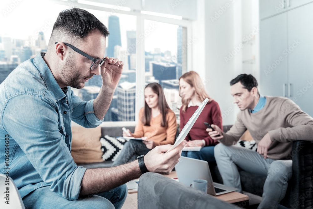 What is this. Interested handsome bespectacled man sitting in the room near his friends overlooking the document and supporting his glasses by his hand. What is this. Interested handsome bespectacled man sitting in the room near his friends overlooking the document and supporting his glasses by his hand.