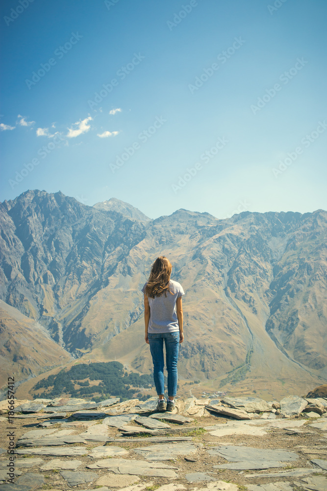 A young girl stands on the edge of a cliff. Mountain in the background. Soldiers stand. Blue sky. Young in jeans and a t-shirt