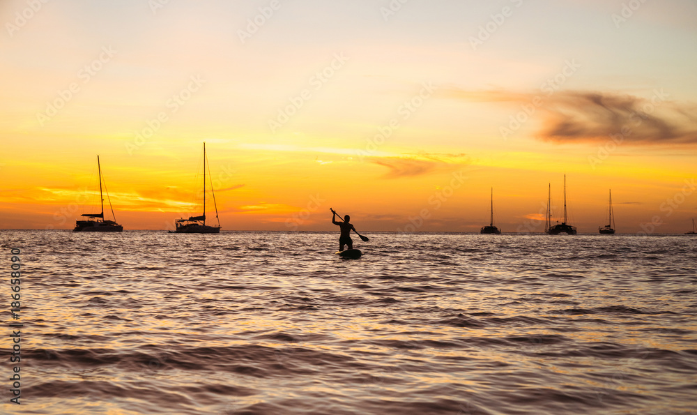 Silhouette of a man with an oar on a swimming board against the background of a sunset and an ocean with yachts. Silhouette of a man with an oar on a swimming board against the background of a sunset and an ocean with yachts.