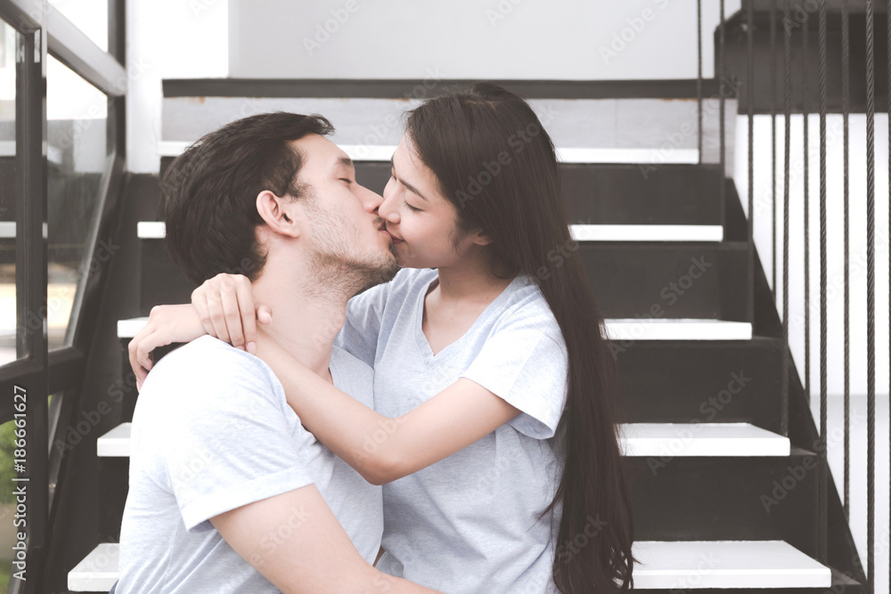 Mixed race lovers concept. Young white male kissing his pretty young  chinese girlfriend on a stairs. Wearing blue jeans, blue shirt and in their  early twenties. Taken indoor. Stock Photo | Adobe