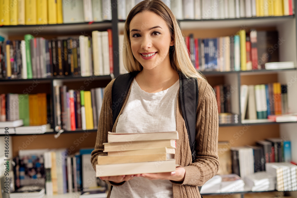 Close up of cute pretty smiling high school student girl standing with a backpack in the library while holding book stack in hands. Close up of cute pretty smiling high school student girl standing with a backpack in the library while holding book stack in hands.