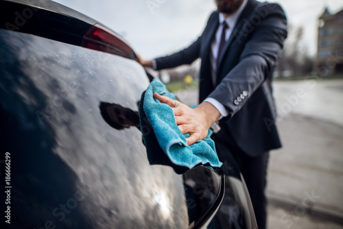 Close up focus hand view of stylish bearded handsome young hardworking man in suit cleaning window with a blue microfiber cloth on the manual self-service car washing station.
