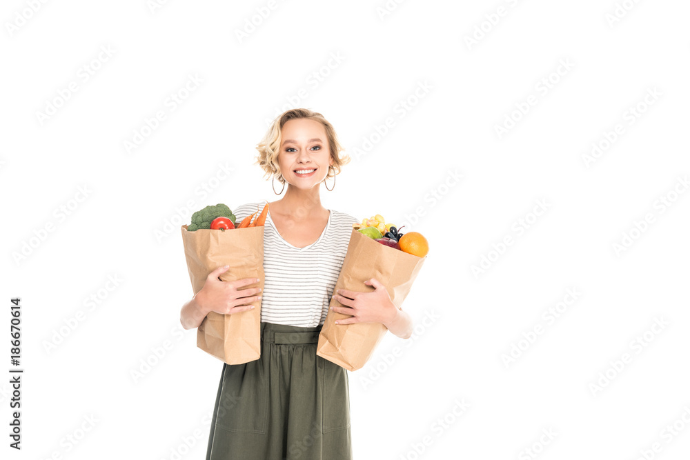 young woman holding paper bags with fruits and vegetables and smiling at camera isolated on white