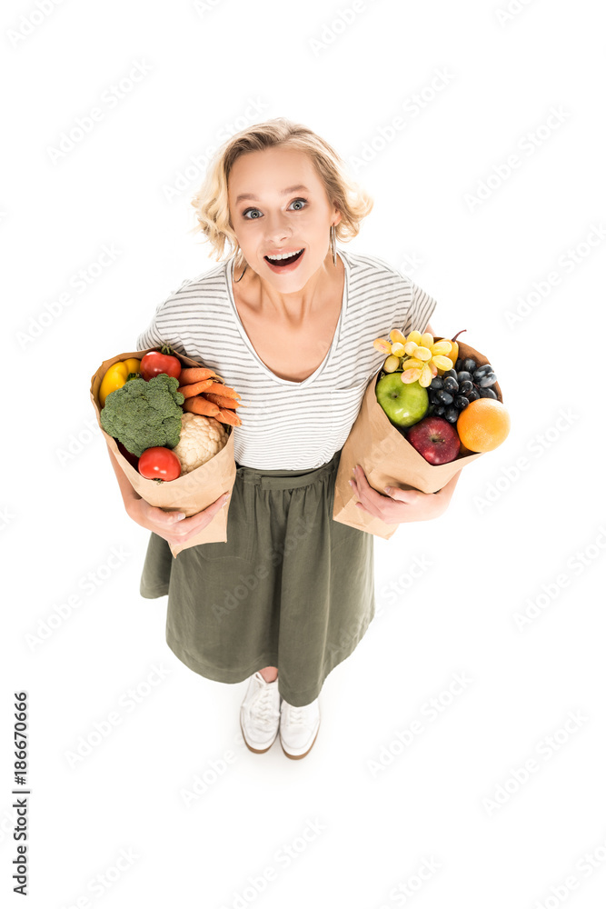 high angle view of cheerful young woman smiling at camera while standing with paper bags full of fresh fruits and vegetables isolated on white high angle view of cheerful young woman smiling at camera while standing with paper bags full of fresh fruits and vegetables isolated on white