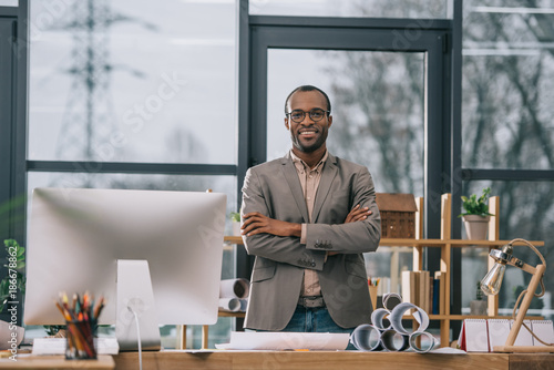 smiling african american architect with crossed arms standing at workplace with computer and blueprints