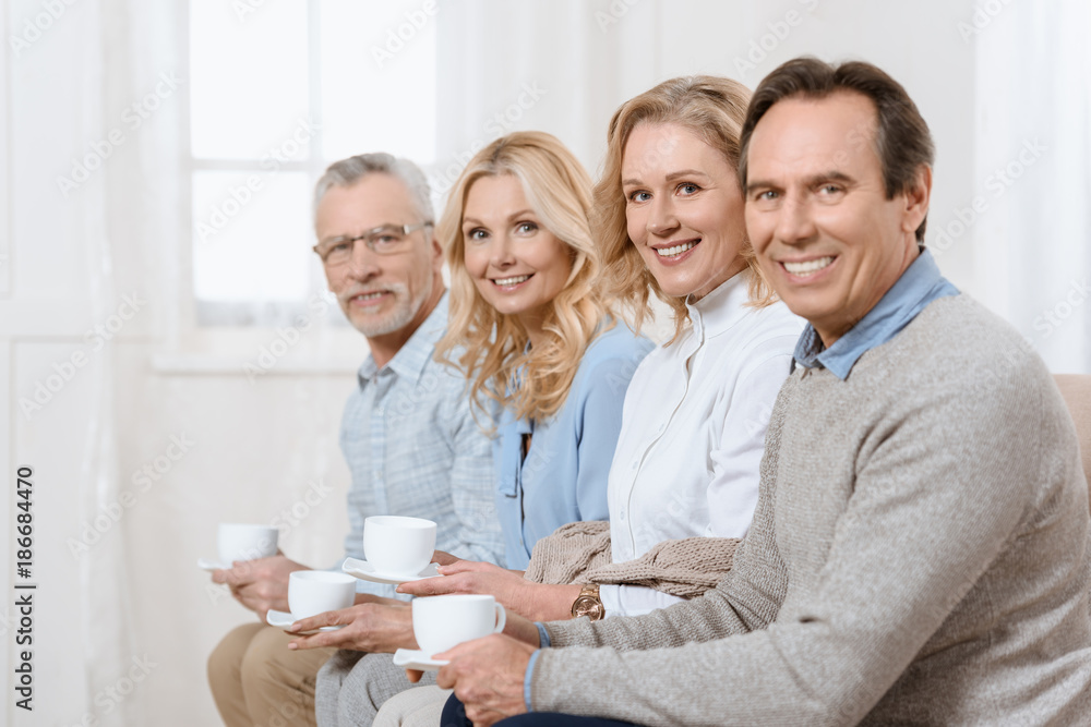 Middle aged men and women having a conversation while drinking tea on sofa looking at camera Middle aged men and women having a conversation while drinking tea on sofa looking at camera