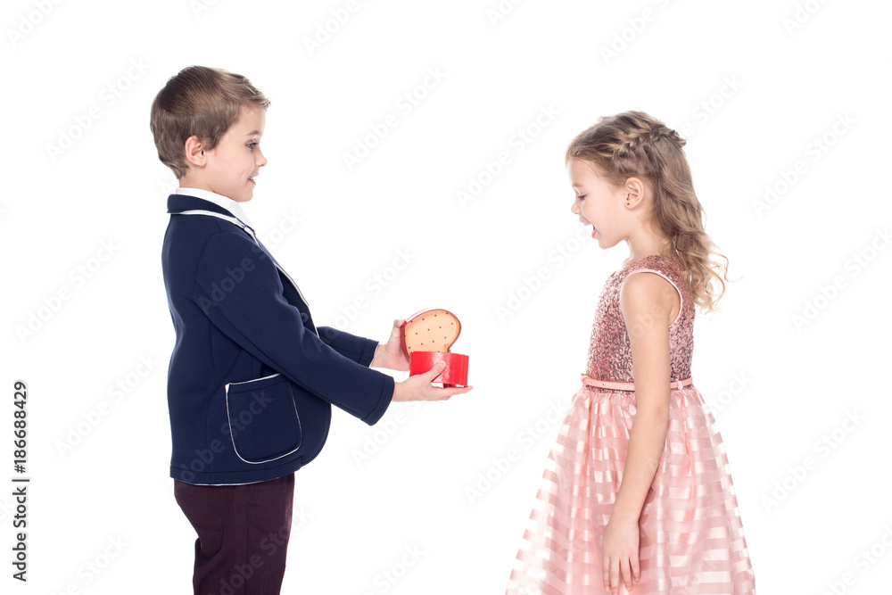 side view of stylish boy opening and presenting heart shaped gift box to surprised little girl isolated on white side view of stylish boy opening and presenting heart shaped gift box to surprised little girl isolated on white