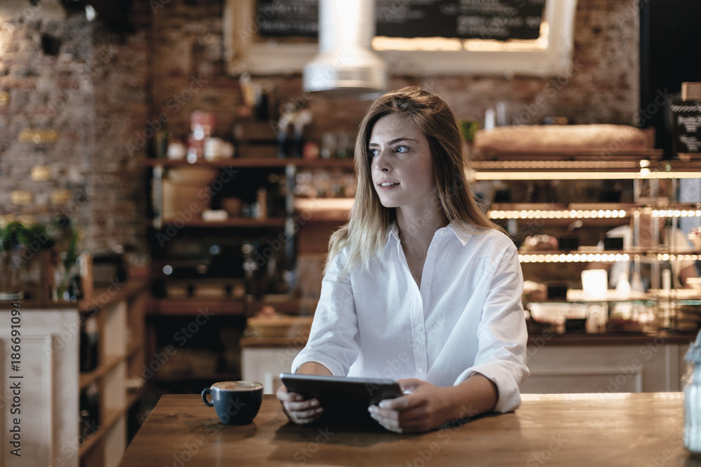 Blond woman sitting in cafe, using tablet, thinking