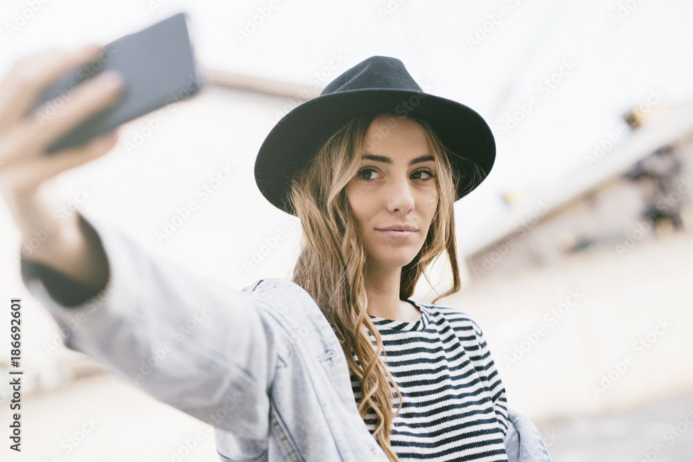 Portrait of fashionable young woman wearing hat taking selfie with smartphone Portrait of fashionable young woman wearing hat taking selfie with smartphone