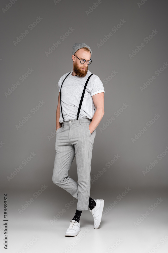 handsome bearded man in suspenders and spectacles standing with hands in  pockets and looking down on grey Stock Photo | Adobe Stock