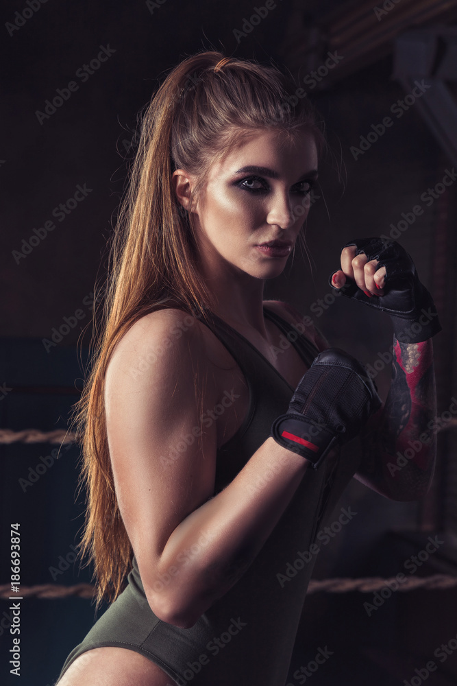 Young pretty boxer woman standing on ring. Female fighter in black glove training in dark gym Young pretty boxer woman standing on ring. Female fighter in black glove training in dark gym