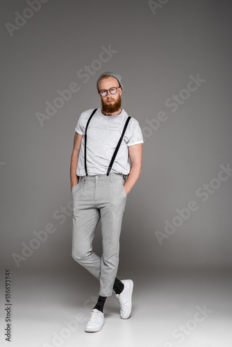 stylish bearded man in hat and spectacles standing with hands in pockets and looking at camera on grey