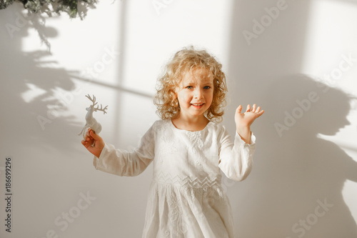 Playful cheerful little beautiful girl with blonde curly hair plays with toy deer, dressed in festive white dress, ready to recieve Christmas presents, isolated over white background with shadow. photo
