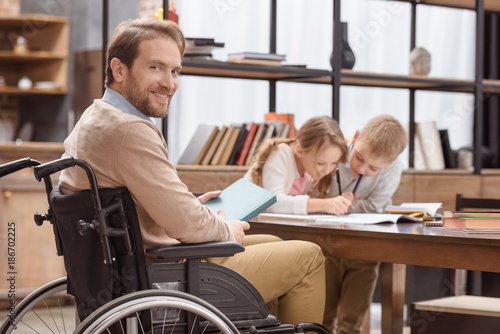 smiling father on wheelchair teaching children at home and looking at camera © LIGHTFIELD STUDIOS