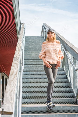 Eastern European Woman wearing bright knitted, off shoulder, loose sweater, black jeans, sneakers, leather cap, walking down stairs in New York, in sunny spring day, looking forward. .