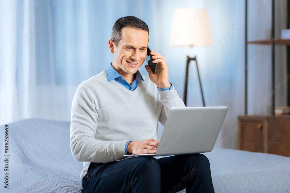 Pleasant chat. Handsome young man sitting on the bed, working on the laptop and talking on the phone while smiling Pleasant chat. Handsome young man sitting on the bed, working on the laptop and talking on the phone while smiling