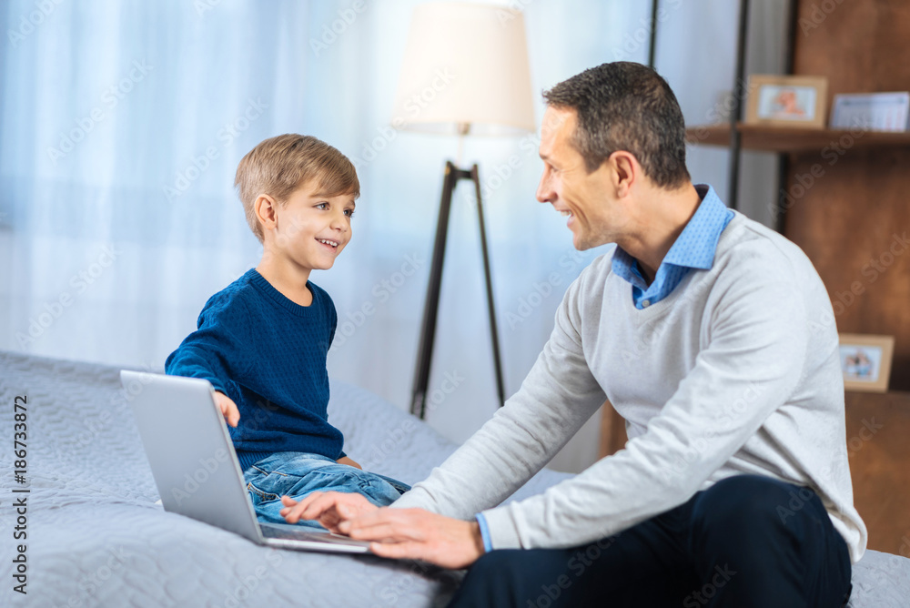 Lively discussion. Happy little boy sitting on the bed next to his father working on the laptop and talking to him, discussing an interesting topic Lively discussion. Happy little boy sitting on the bed next to his father working on the laptop and talking to him, discussing an interesting topic