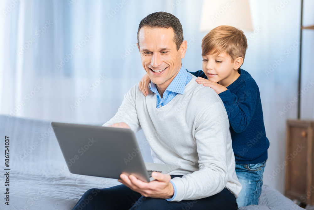 Childlike curiosity. Pleasant upbeat man sitting on the bed and pointing at the laptop, explaining his curious little son the basics of his work while the boy back-hugging him Childlike curiosity. Pleasant upbeat man sitting on the bed and pointing at the laptop, explaining his curious little son the basics of his work while the boy back-hugging him